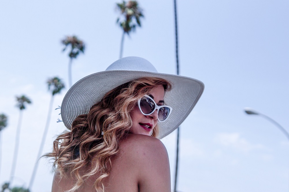 Femeie blondă în vacanță, cu bucle, pălărie pe cap și ochelari de soare.